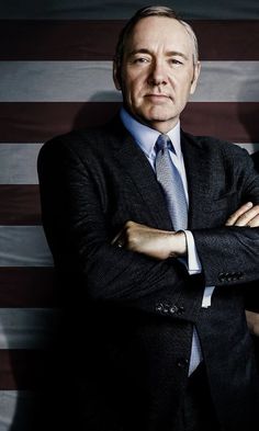 a man wearing a suit and tie standing in front of an american flag with his arms crossed