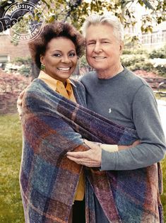 an older man and woman hugging each other in front of a tree with their arms wrapped around them