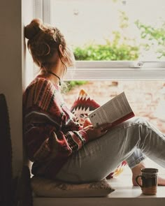 a woman sitting on a window sill reading a book and holding a coffee mug