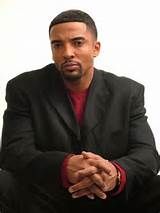 a man in a black suit sitting down with his hands folded on his knees and looking at the camera