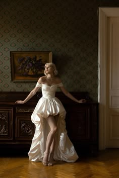 a woman in a white dress is standing on a wooden floor next to a dresser