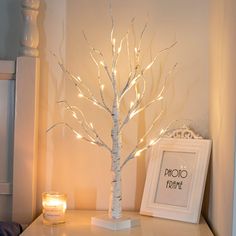 a small white tree with lights on it next to a candle holder and framed photograph