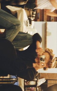 a woman holding a camera in front of her face while sitting on a chair with other people behind her