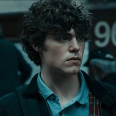 a young man with curly hair wearing a blue shirt and black jacket looking at the camera