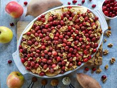 an apple and cranberry dessert in a white dish surrounded by apples, pears, and other fruits