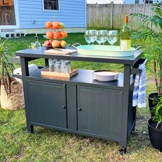 an outdoor bar cart with wine glasses and fruit on it in the grass next to a blue house