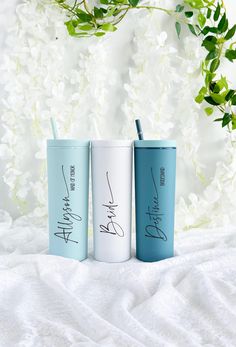 three different colored tumblers with writing on them sitting on a white blanket next to flowers