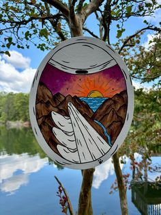 a painting hanging from the side of a tree next to a body of water with mountains in the background