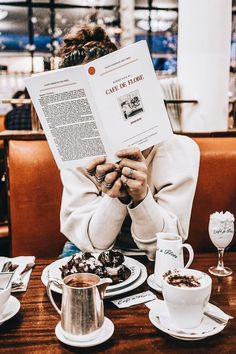 a woman reading a book while sitting at a table with cups and saucers on it