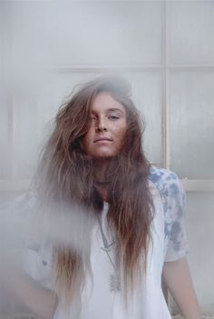 a woman with long hair standing in front of a white wall and looking at the camera