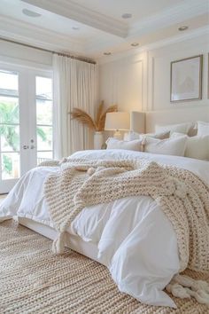 a bed with white linens and blankets in a bedroom next to two large windows