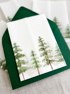 three folded cards with trees on them sitting on a table next to green envelopes