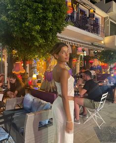 a woman in a white dress is standing on the sidewalk near some tables and chairs