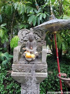 a statue with fruit on it in the middle of some bushes and trees, next to an umbrella