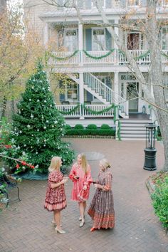 three women in dresses walking down a brick walkway near christmas trees and a large white house