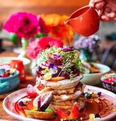 a person pouring syrup on top of a stack of pancakes covered in fruit and toppings