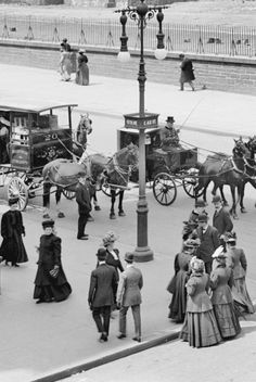 an old black and white photo of people on the street with horse drawn carriages behind them