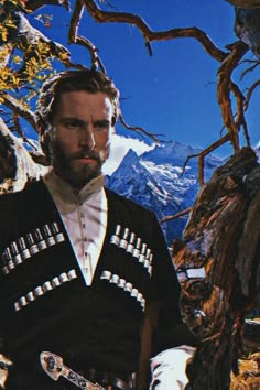 a man with a beard is standing in front of a tree and mountains, holding a snowboard