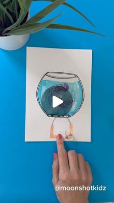 someone is drawing a fish in a bowl with water and glue on the paper, next to a potted plant