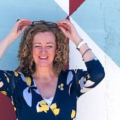 a woman with curly hair is smiling and holding her hands on her head while standing in front of a wall