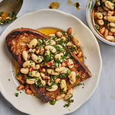 a white plate topped with meat and beans next to a bowl of vegetables on top of a table