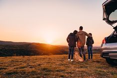 three people and a dog standing in front of a car with the sun setting behind them