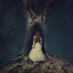 a woman in a white dress standing next to a large tree with its roots exposed