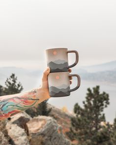 a person holding two coffee mugs in their hands on top of a mountain overlooking the ocean