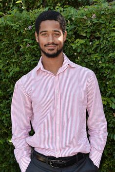 a man standing in front of bushes wearing a red and white striped shirt with his hands in his pockets