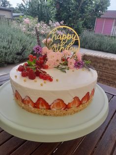 a white cake with strawberries and flowers on top sitting on a wooden table outside