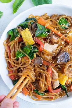 someone holding chopsticks over a bowl of noodles with vegetables and meat on it