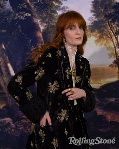 a woman standing in front of a painting wearing a black coat with gold flowers on it