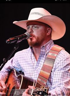 a man wearing a cowboy hat and holding a guitar in front of a microphone on stage