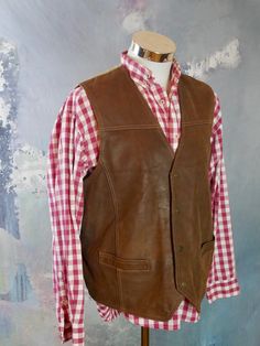 This 1990s brown leather vest has a V neckline, and closes in the front with four snap buttons. The vintage pointed-front Western waistcoat has two pockets on the front, and features topstitching on the leather. The vest has a full beige canvas (cotton) back with a leather half-belt with buckle. Chest = Up to 48 inches (121.92cm) Vest Length (front) = 26 inches (66.04cm) Vest Length (back) = 24 inches (60.96cm) Brand label: Pionier Sportive Size: 46 to 48 US/UK Material: Genuine Leather --- COND Rugged Brown Vest Outerwear, Brown Rugged Vest For Winter, Rugged Brown Winter Vest, Brown Rugged Winter Vest, Brown Sleeveless Vest For Western-themed Events, Vintage Leather Vest Outerwear, Western Leather Vest For Fall, Retro Brown Vest For Workwear, Retro Brown Vest For Work