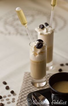 two glasses filled with drinks on top of a table next to spoons and coffee beans