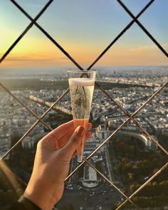 someone holding up a glass in front of a fence overlooking the eiffel tower