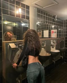 a woman standing in front of a bathroom mirror next to a urinal and sink