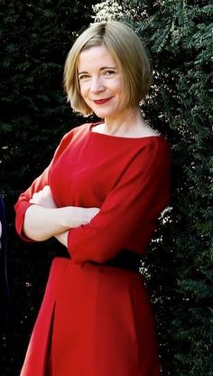 a woman in a red dress standing next to a bush with her arms crossed and smiling at the camera