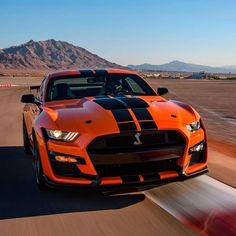 an orange and black mustang driving down a road with mountains in the background at sunset