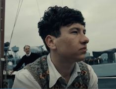 a man in a vest and tie sitting on a boat with other men behind him