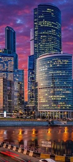 the city skyline is lit up at night with cars passing by in front of it