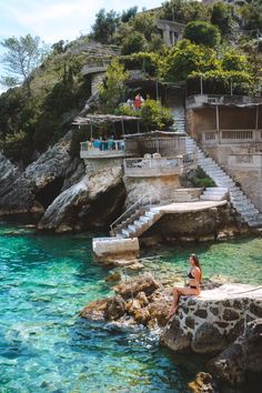 a woman sitting on the edge of a cliff next to blue water and steps leading up to a house