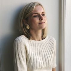 a woman leaning against a wall with her head turned to the side, wearing a white sweater