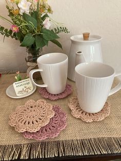 crocheted coasters and coffee cups on a table with flowers in the background