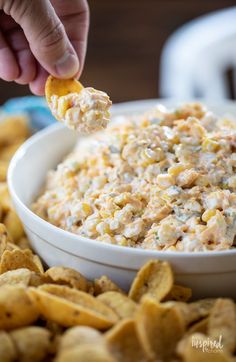 a person dipping corn into a bowl of dip with crackers on the side and chips scattered around