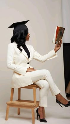 a woman in a white suit and black hat sitting on a chair holding a book