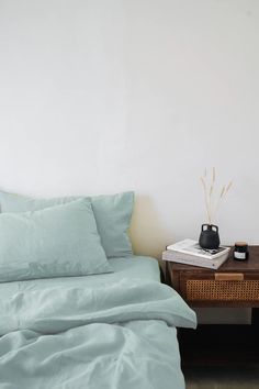 a bed with green sheets and pillows next to a night stand on a wooden table