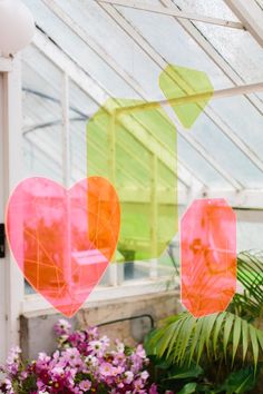 some pink and green hearts hanging from a glass ceiling in a greenhouse with purple flowers