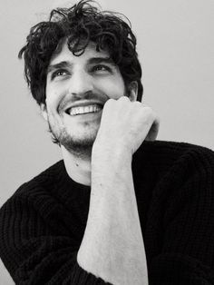 a black and white photo of a man with curly hair smiling at the camera while holding his hand to his face