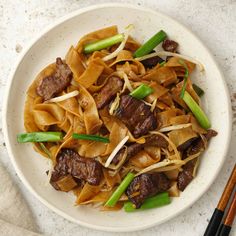 a white plate topped with beef and noodles next to chopsticks on a table